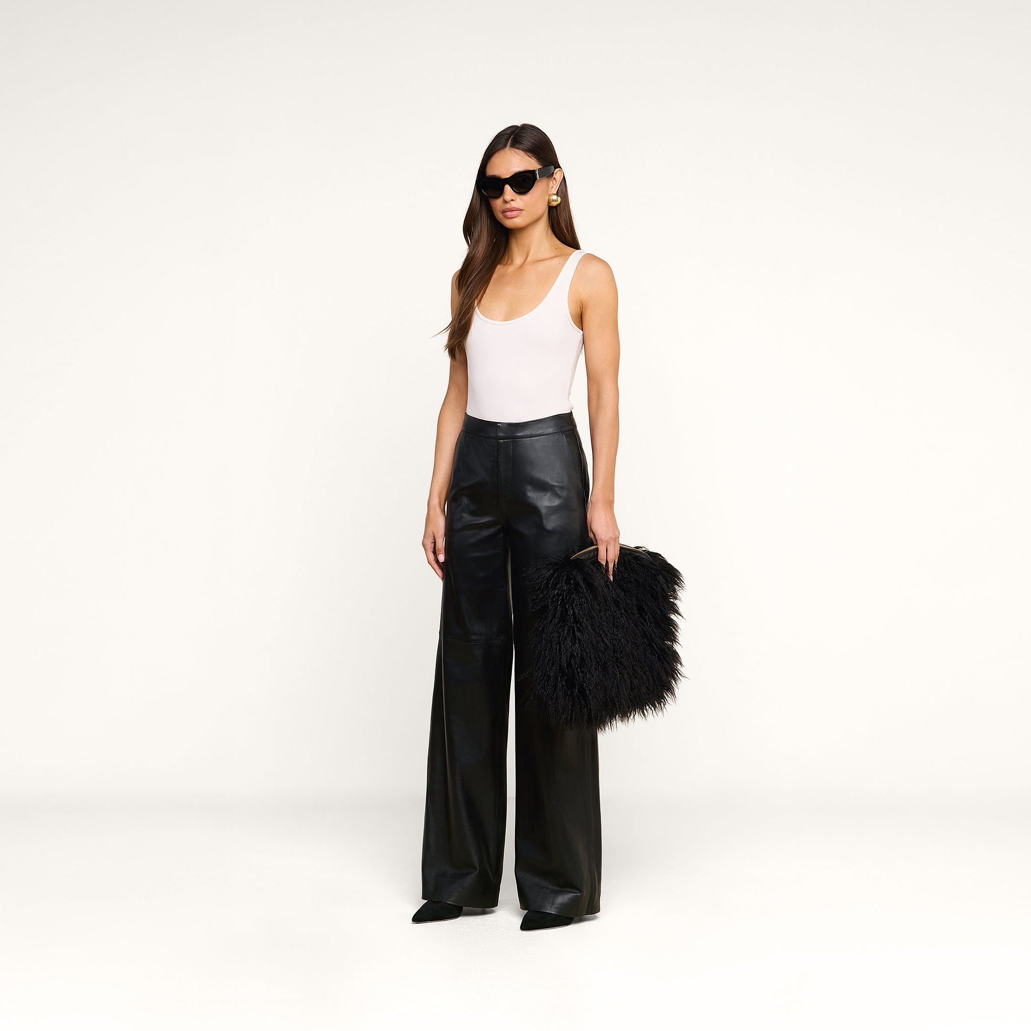 A woman stands against a plain white background wearing a white tank top, black wide-leg leather pants, black pointed shoes, and sunglasses, holding the Adelina Faux Fur Clutch.