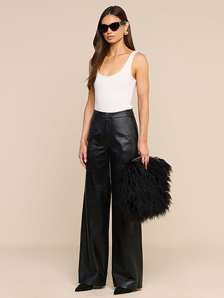 Woman in sunglasses wears a white tank top, black wide-leg leather pants, and black pointed shoes. She holds the Adelina Faux Fur Clutch—a true statement accessory—and stands against a beige background.
