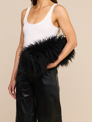 A woman wears a white tank top and black leather pants, holding the Adelina Faux Fur Clutch—a plush black statement accessory—under her arm. The background is plain beige.