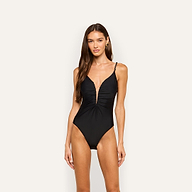 A woman stands facing forward against a plain light background, wearing the Lilou One-Piece Swimsuit—a black one-piece featuring a deep V-neckline, high-cut legs, and gold chain hardware strap details.