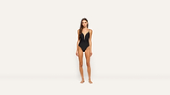 A woman stands barefoot against a plain light background, wearing a black one-piece swimsuit with thin straps. The swimsuit’s classic design highlights her elegant silhouette.