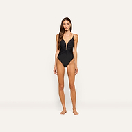A woman stands barefoot against a plain light background, wearing the Lilou One-Piece Swimsuit, which features a black color, ruched front, thin shoulder straps, and high cut legs.