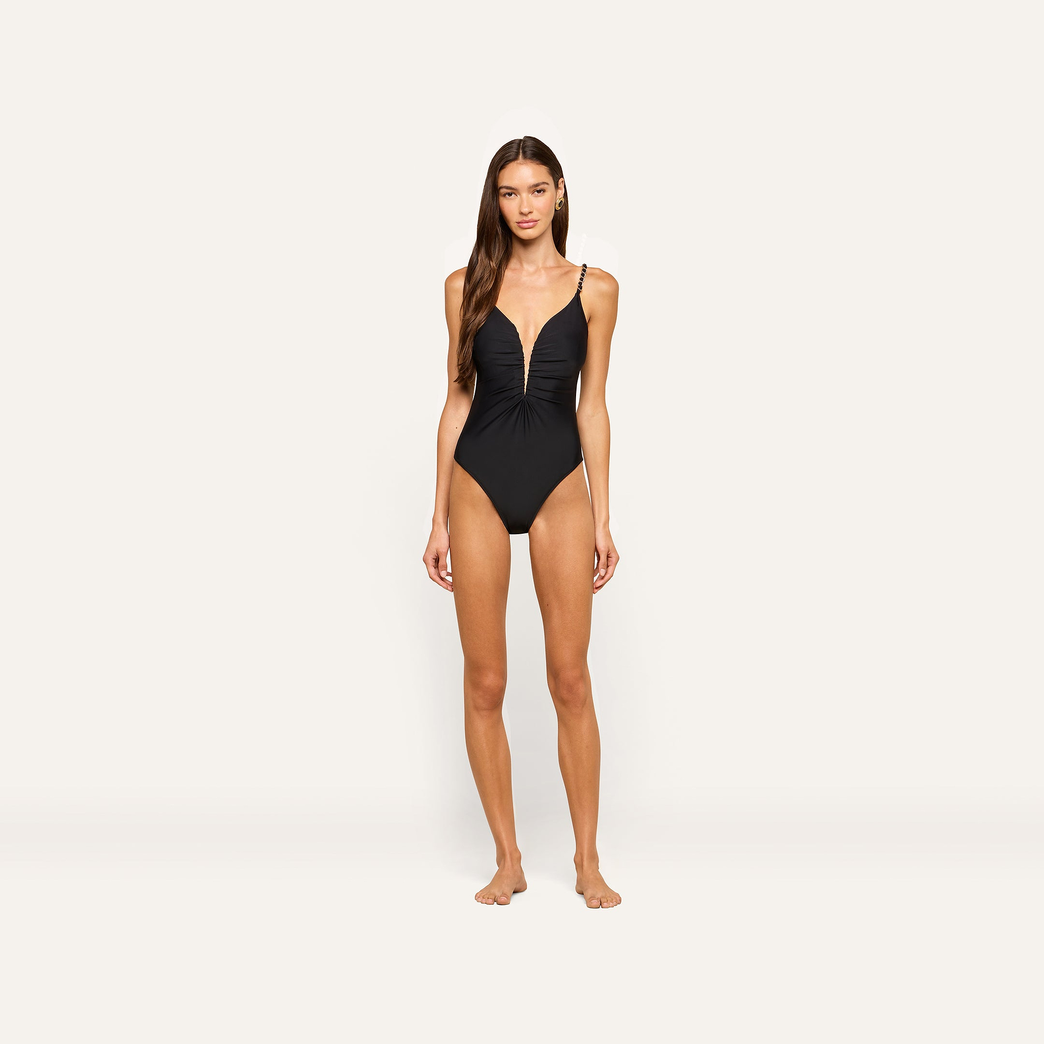 A woman stands barefoot against a plain light background, wearing the Lilou One-Piece Swimsuit, which features a black color, ruched front, thin shoulder straps, and high cut legs.