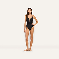 A woman stands barefoot against a plain light background, wearing the Lilou One-Piece Swimsuit—a high cut black one-piece with thin straps, gold chain hardware, and ruched detailing on the front.