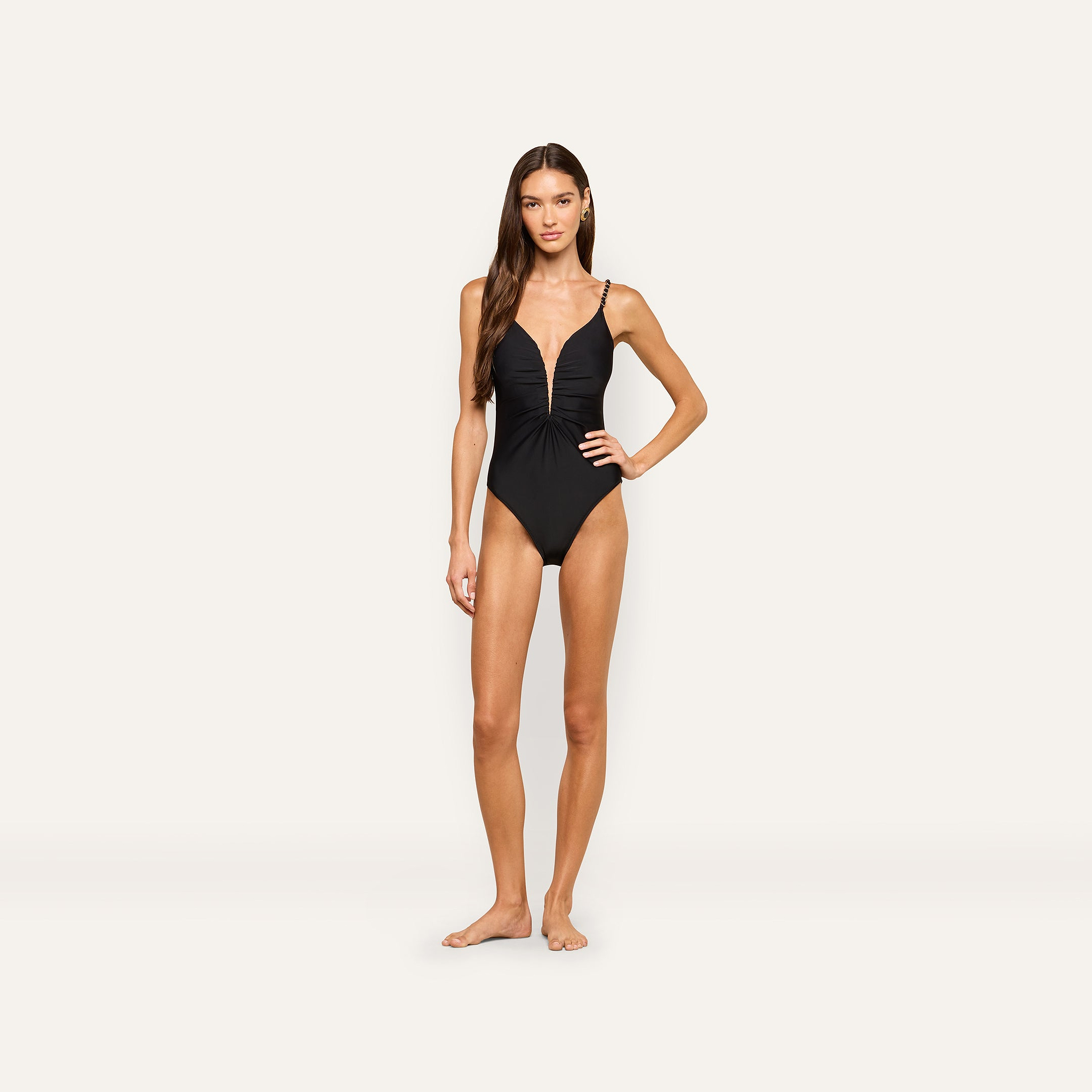 A woman stands barefoot against a plain light background, wearing the Lilou One-Piece Swimsuit—a high cut black one-piece with thin straps, gold chain hardware, and ruched detailing on the front.
