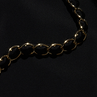 The Lilou One-Piece Swimsuit features a shiny gold chain with large, rounded links, reminiscent of gold chain hardware, elegantly displayed diagonally across a smooth black fabric background.