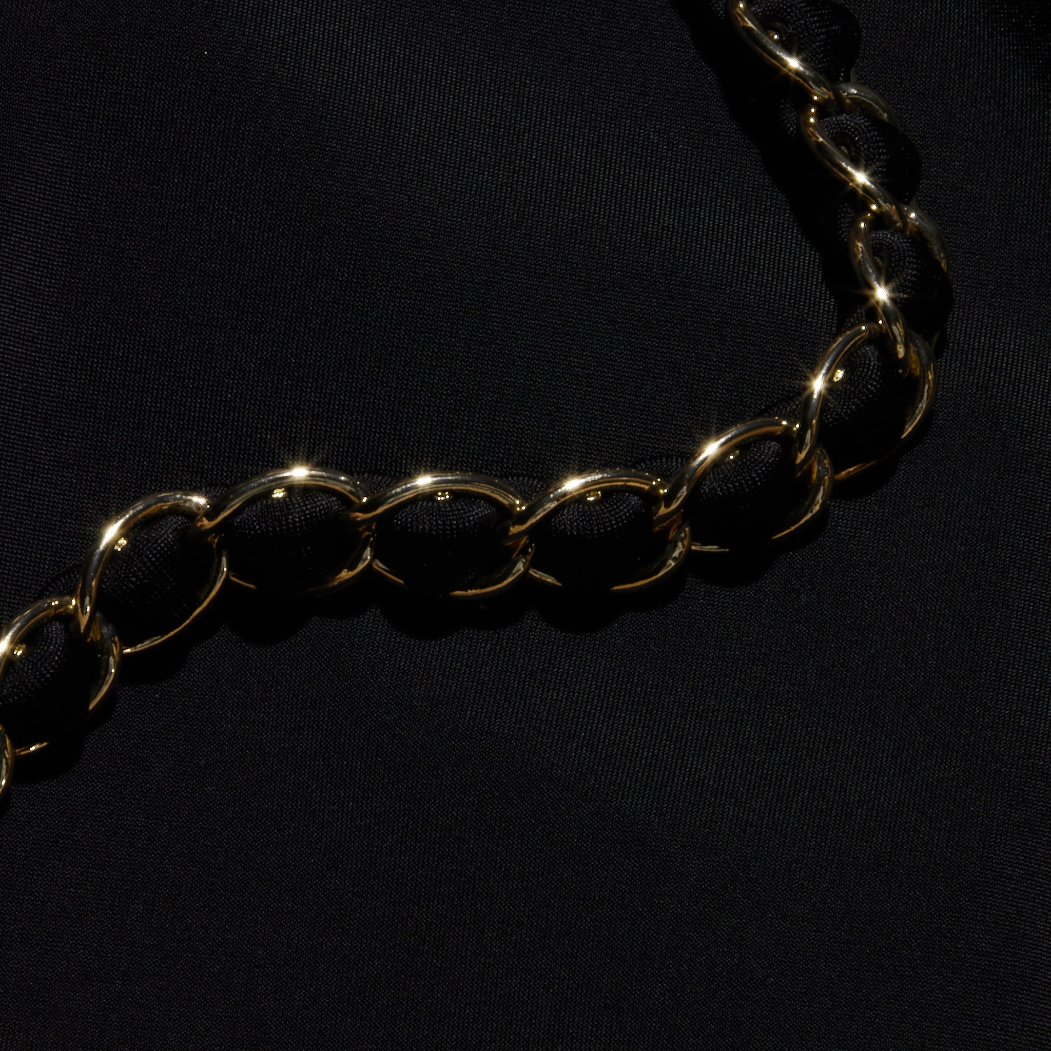 The Lilou One-Piece Swimsuit features a shiny gold chain with large, rounded links, reminiscent of gold chain hardware, elegantly displayed diagonally across a smooth black fabric background.