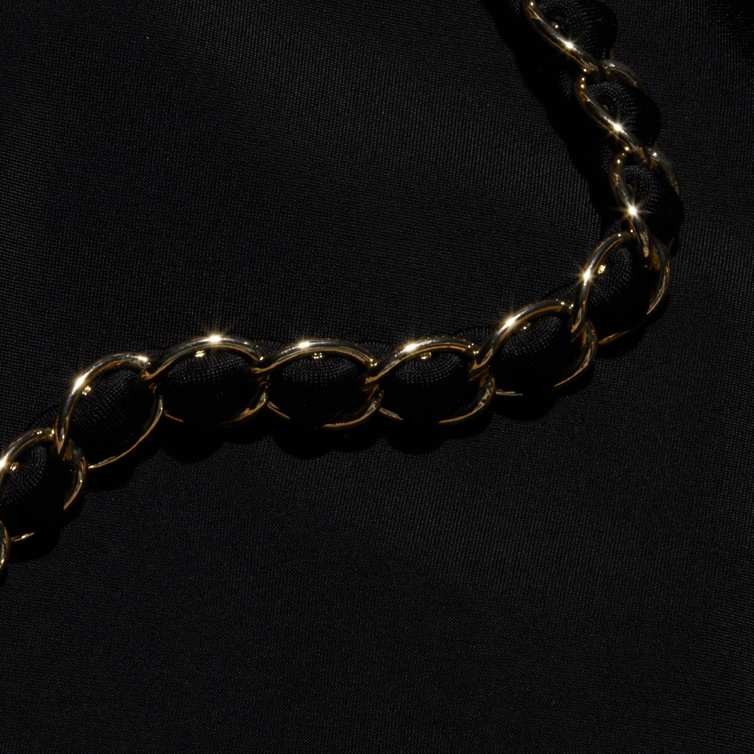The Lilou One-Piece Swimsuit features a shiny gold chain with large, rounded links, reminiscent of gold chain hardware, elegantly displayed diagonally across a smooth black fabric background.