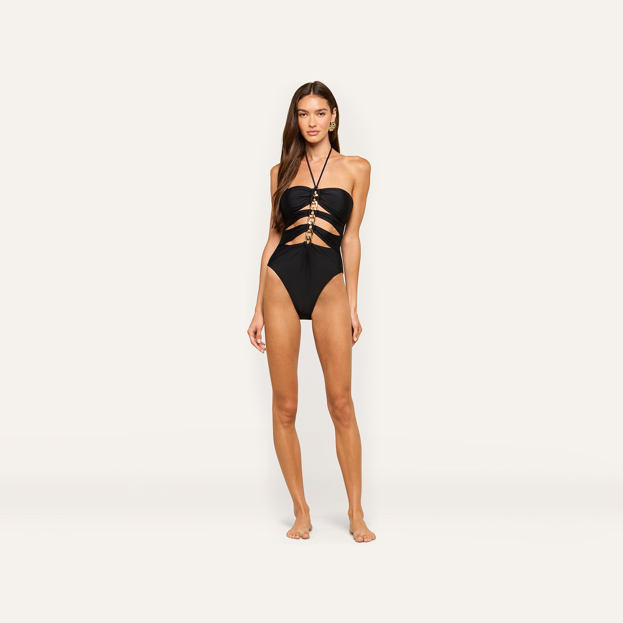 A woman stands barefoot wearing the Opus Halter One-Piece Swimsuit, a black one-piece with cutouts and gold ring accents, as she poses against a plain, light background.