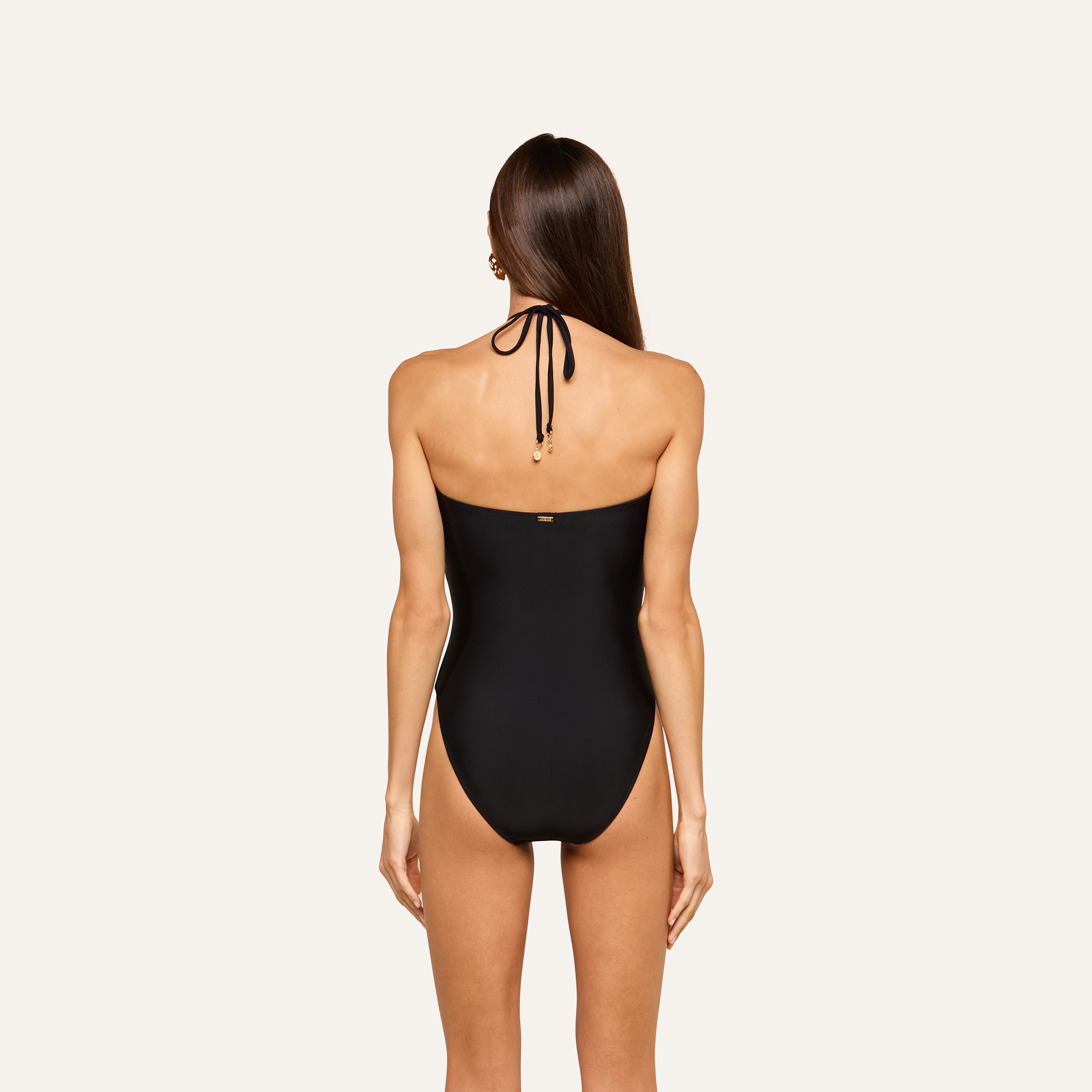 A woman with long brown hair is seen from the back against a plain light background, wearing the Opus Halter One-Piece Swimsuit in black. This chic resort swimsuit effortlessly blends style and coverage for any sun-soaked day.