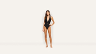 A woman stands barefoot against a plain background, wearing a black belted one-piece swimsuit with a deep V neckline.