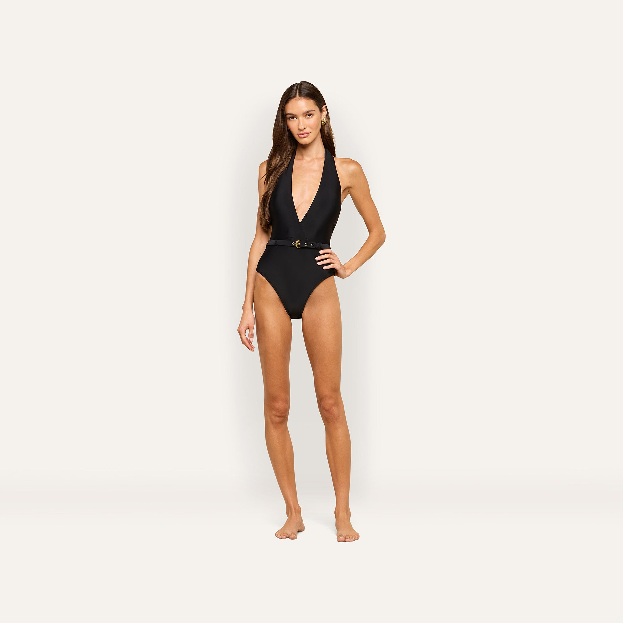 A woman stands barefoot against a plain, light background wearing the Sidney Plunge One-Piece Swimsuit in matte black, featuring a belted waist and convertible tie straps.