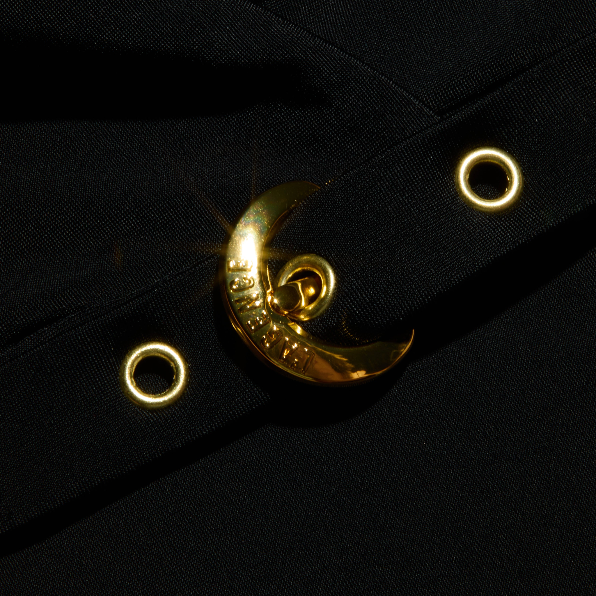 A crescent-shaped, shiny gold buckle is attached to the Sidney Plunge One-Piece Swimsuit’s black fabric strap, which is accented with gold eyelets and photographed against a black background.