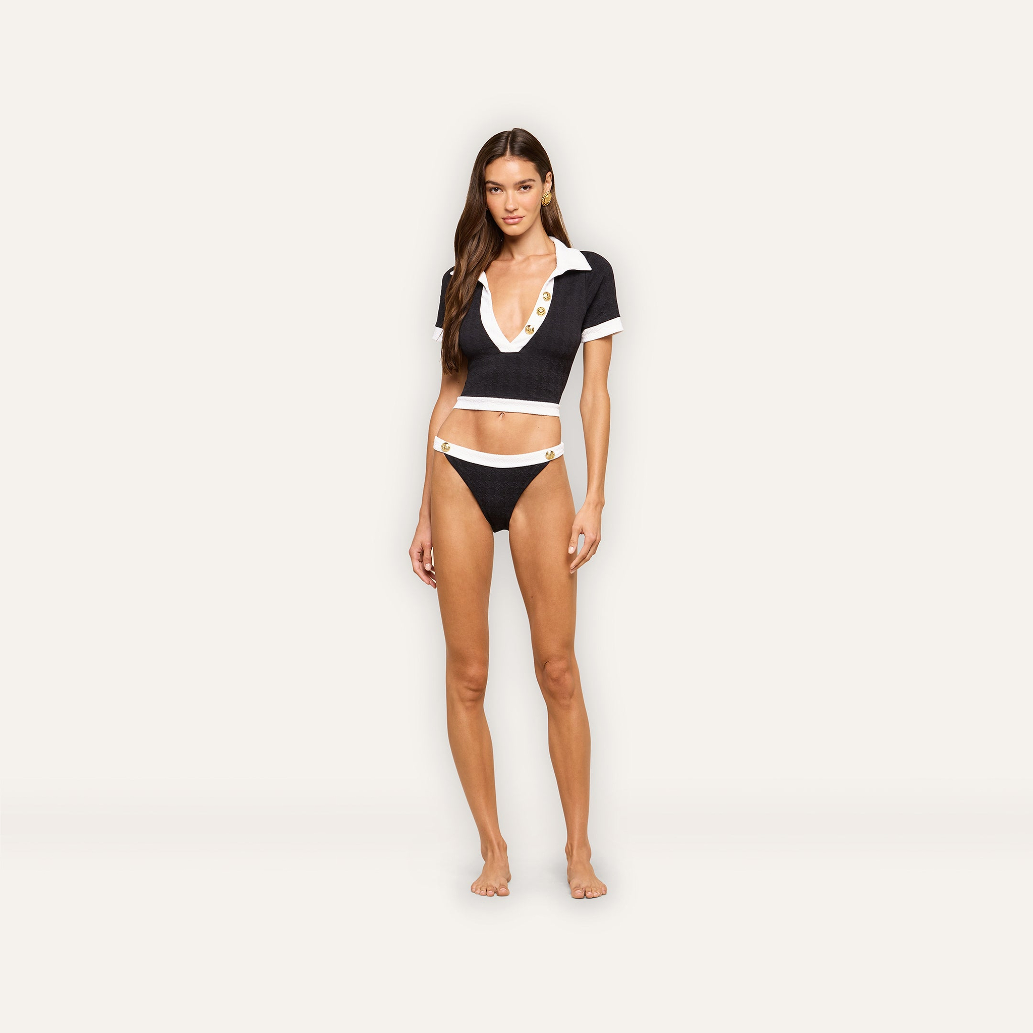 A woman stands against a light background wearing the Laguna Rash Guard, a short-sleeve black crop top styled like a polo with a deep V-neck and white trim, paired with matching bikini bottoms featuring white straps and gold details.