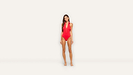A woman stands barefoot against a plain white background, wearing a bold red one-piece swimsuit with a deep V-neck and halter-style straps.