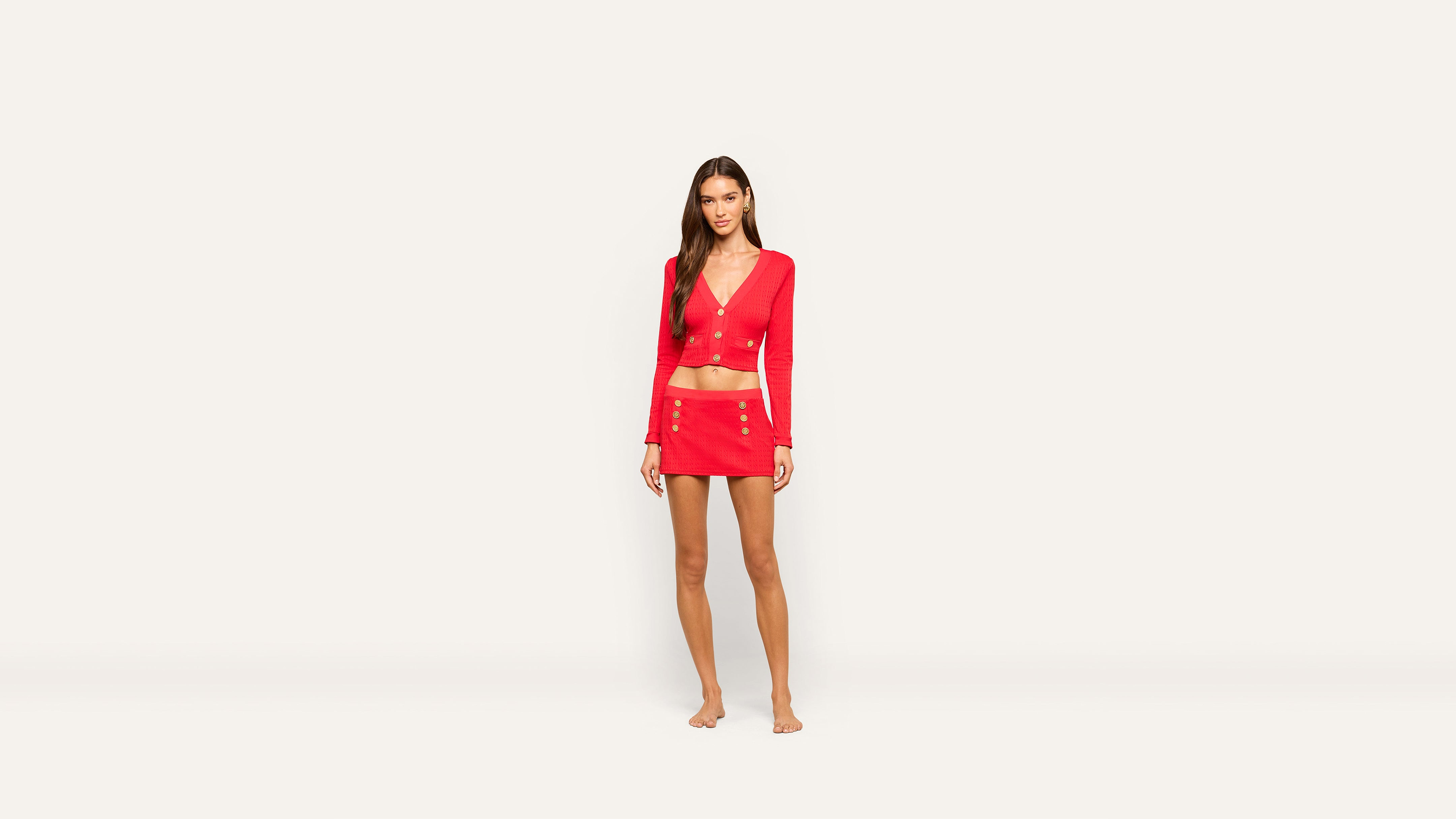 A woman stands barefoot against a plain white background, wearing a bright red long-sleeve crop top and a matching red mini skirt adorned with gold buttons.
