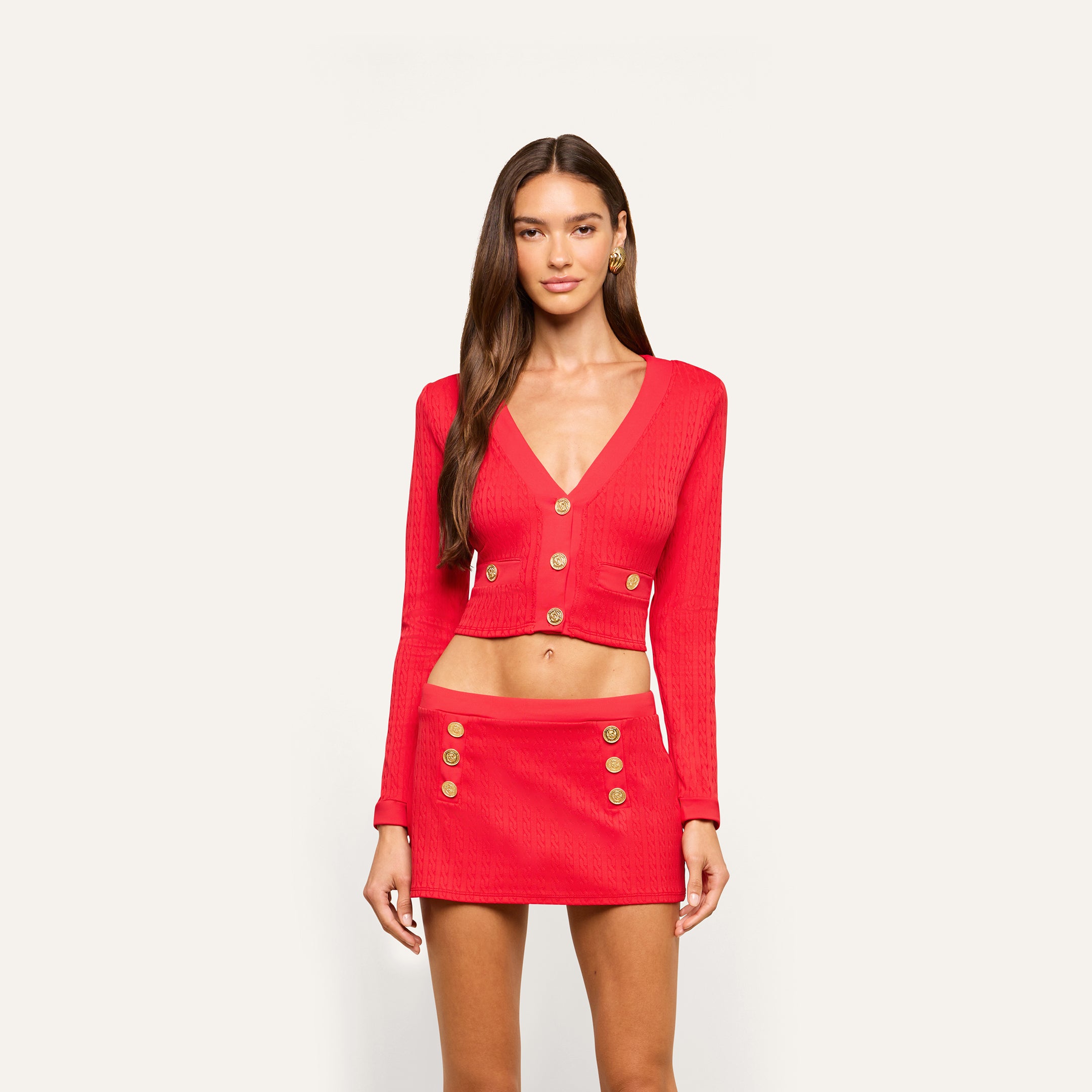 A woman with long brown hair wears a red cropped cardigan with gold buttons and the Coco Mini Skirt Cover-Up, also in red with gold buttons, standing against a plain light background.