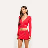 A woman wearing a bright red, long-sleeve, buttoned crop top and the Coco Mini Skirt Cover-Up with gold buttons stands against a plain light background.