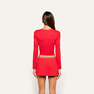 A woman with long brown hair stands facing away, wearing a fitted, long-sleeved red top and the Coco Mini Skirt Cover-Up against a plain light background.