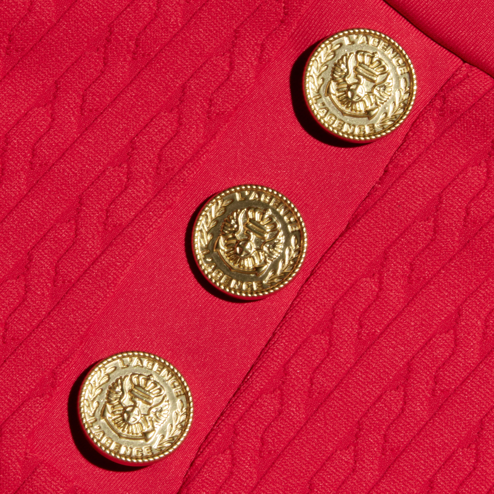 Three ornate gold buttons with embossed designs are sewn onto the textured red fabric with a wavy pattern of the Coco Mini Skirt Cover-Up, making it an elegant detail for your swim skirt or cover-up ensemble.