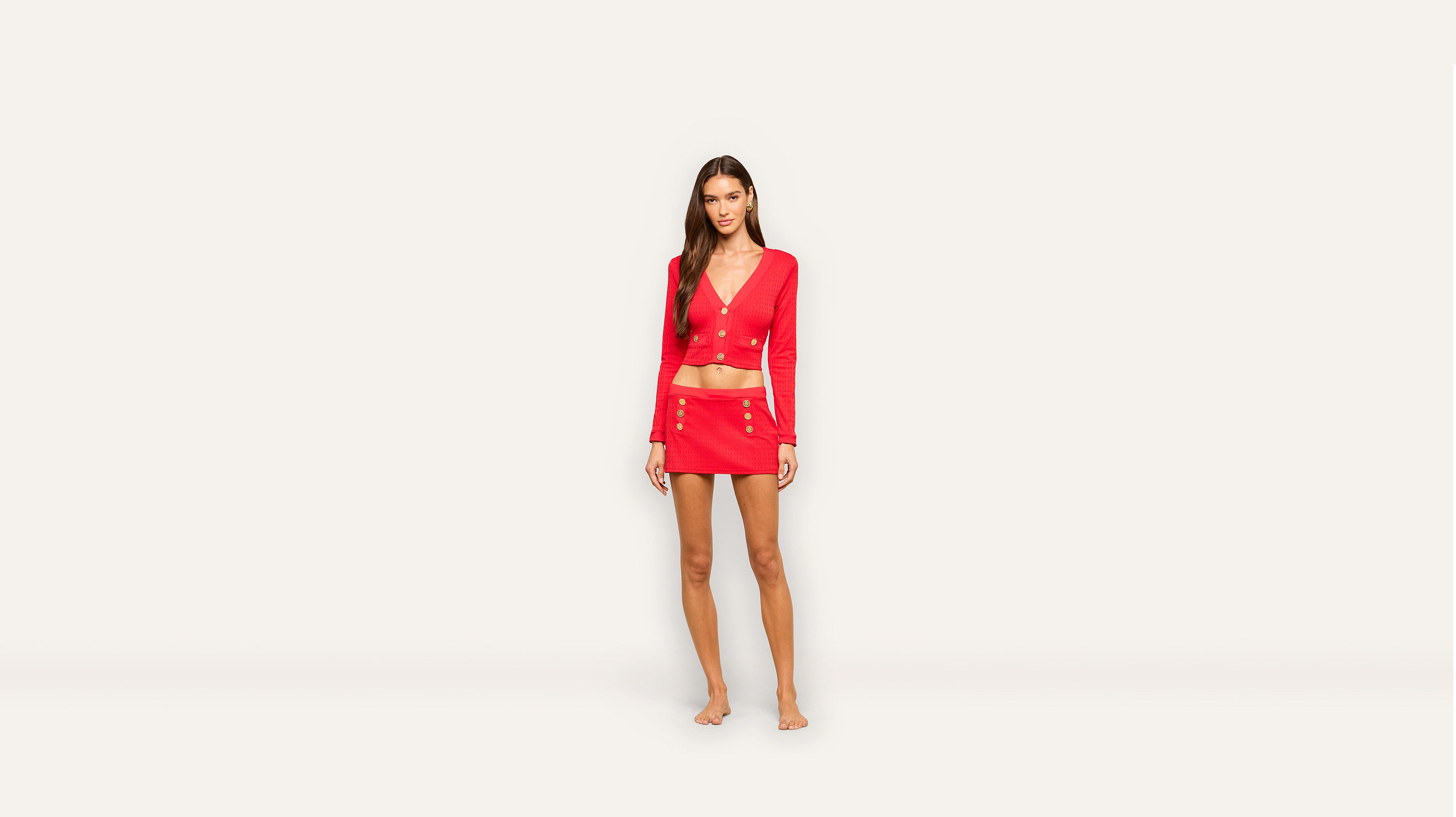 A woman stands barefoot against a plain light background, wearing a vibrant red long-sleeve crop top and matching mini skirt adorned with gold buttons.