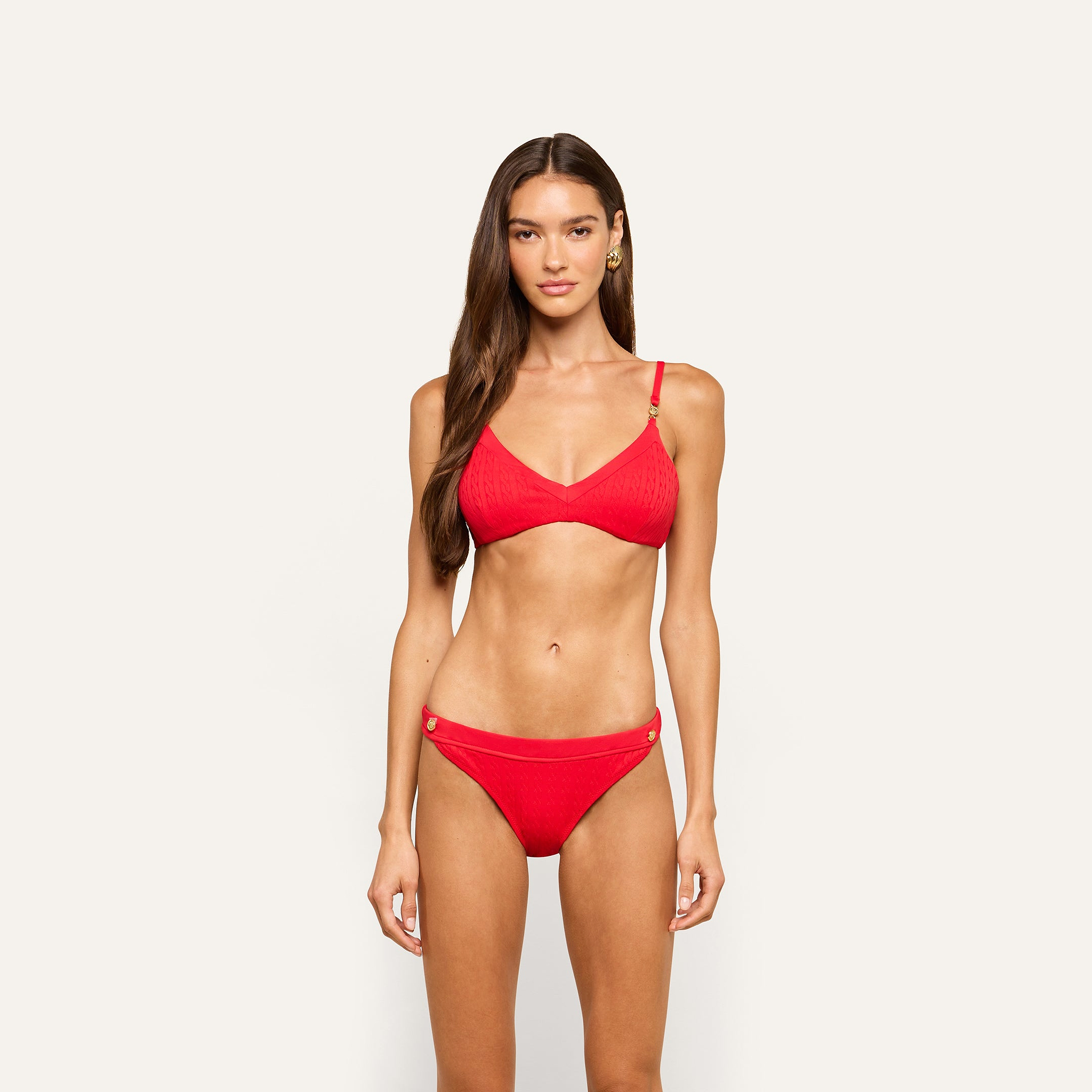 A woman with long brown hair wears the Playa Bikini Top in bright red cable knit, standing against a plain, light background and facing forward with a neutral expression.
