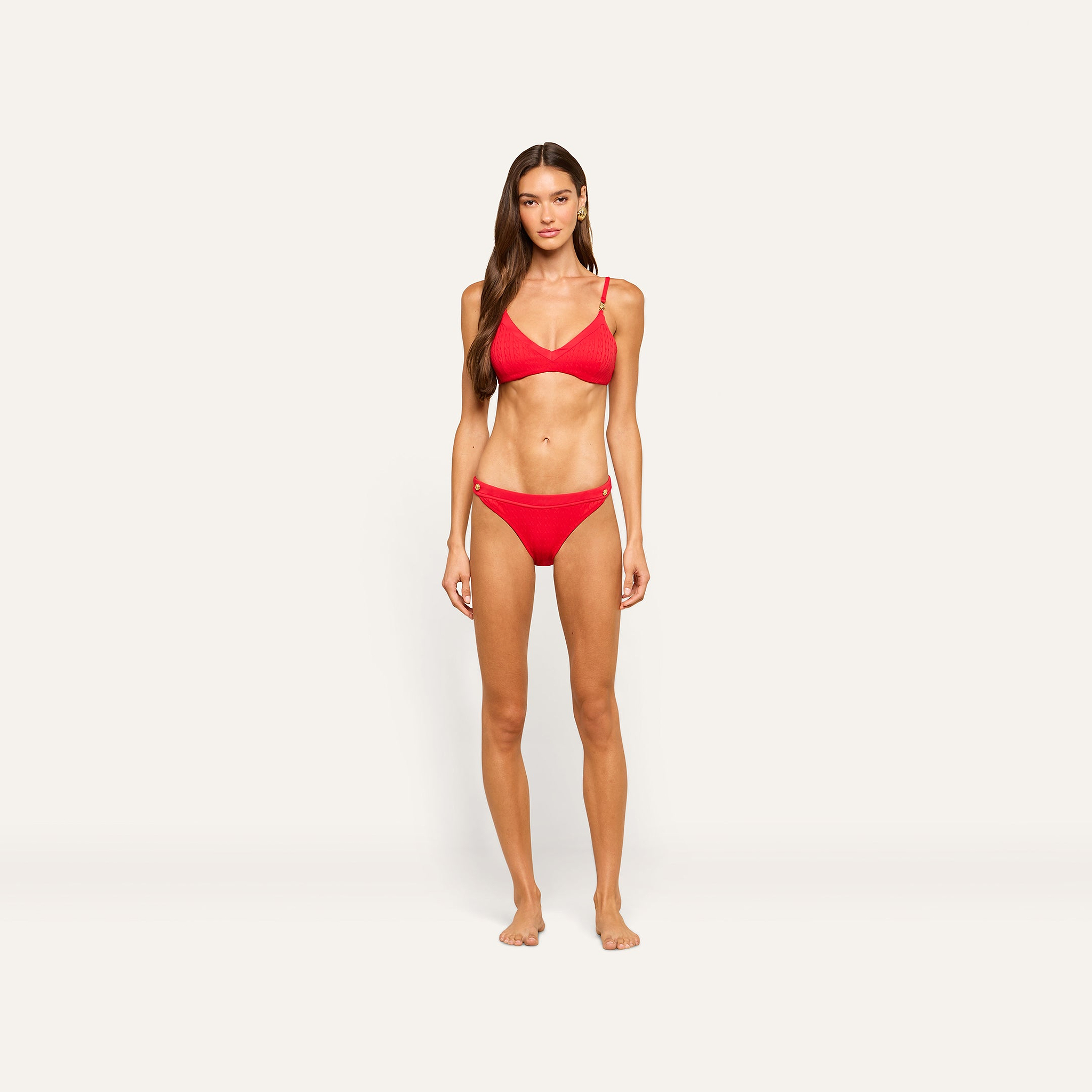 A woman with long brown hair stands against a plain light background, wearing the bright red Playa Bikini Top and no shoes, looking directly at the camera with a neutral expression.