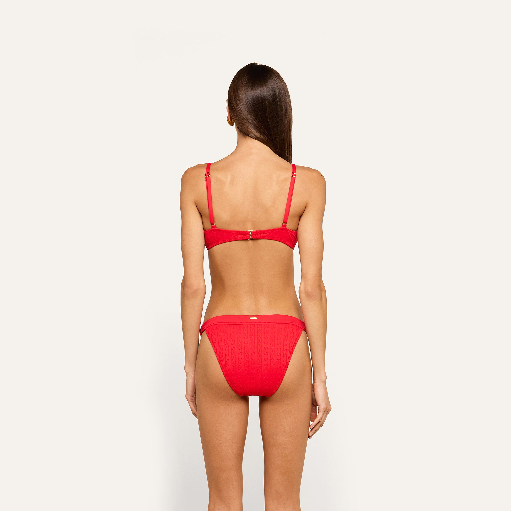 A woman stands with her back to the camera, wearing the bright red Playa Bikini Top, which features adjustable straps and textured cable knit fabric. The background is plain and light-colored.