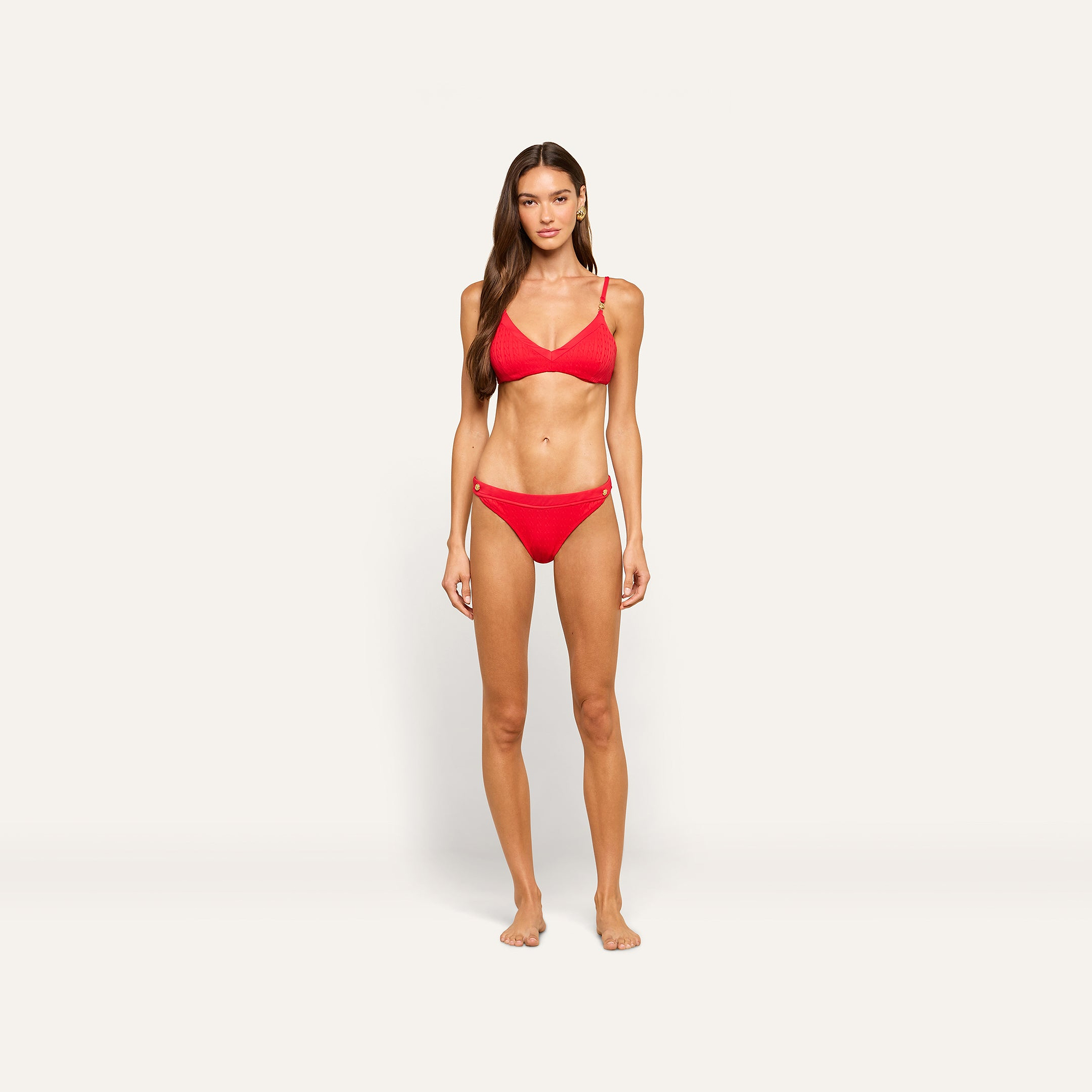 A woman stands facing forward, wearing the Jean Bikini Bottom in red, featuring a high-leg cut and textured swim fabric, against a plain, light-colored background.