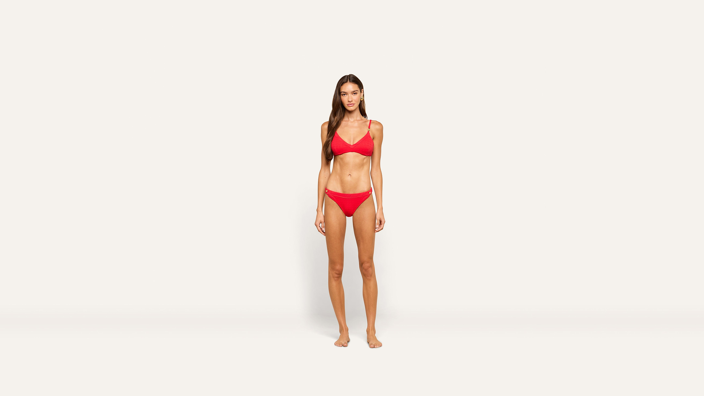 Woman with long brown hair stands barefoot against a plain white background, wearing a red two-piece bikini set. She is facing forward with a neutral expression and relaxed posture.