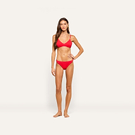 A woman stands against a plain light background, wearing a matching red bikini crafted from textured swim fabric. She has long brown hair, one hand on her hip, and a neutral expression. She is wearing the Jean Bikini Bottom, whose high-leg cut enhances the sleek silhouette of her swimwear.