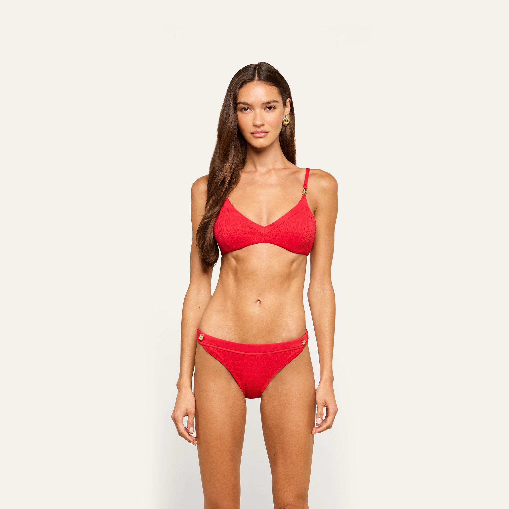 A woman with long brown hair stands facing forward, wearing a bright red Jean Bikini Bottom made from textured swim fabric with thin straps and decorative buttons on the high-leg bikini bottoms, against a plain off-white background.
