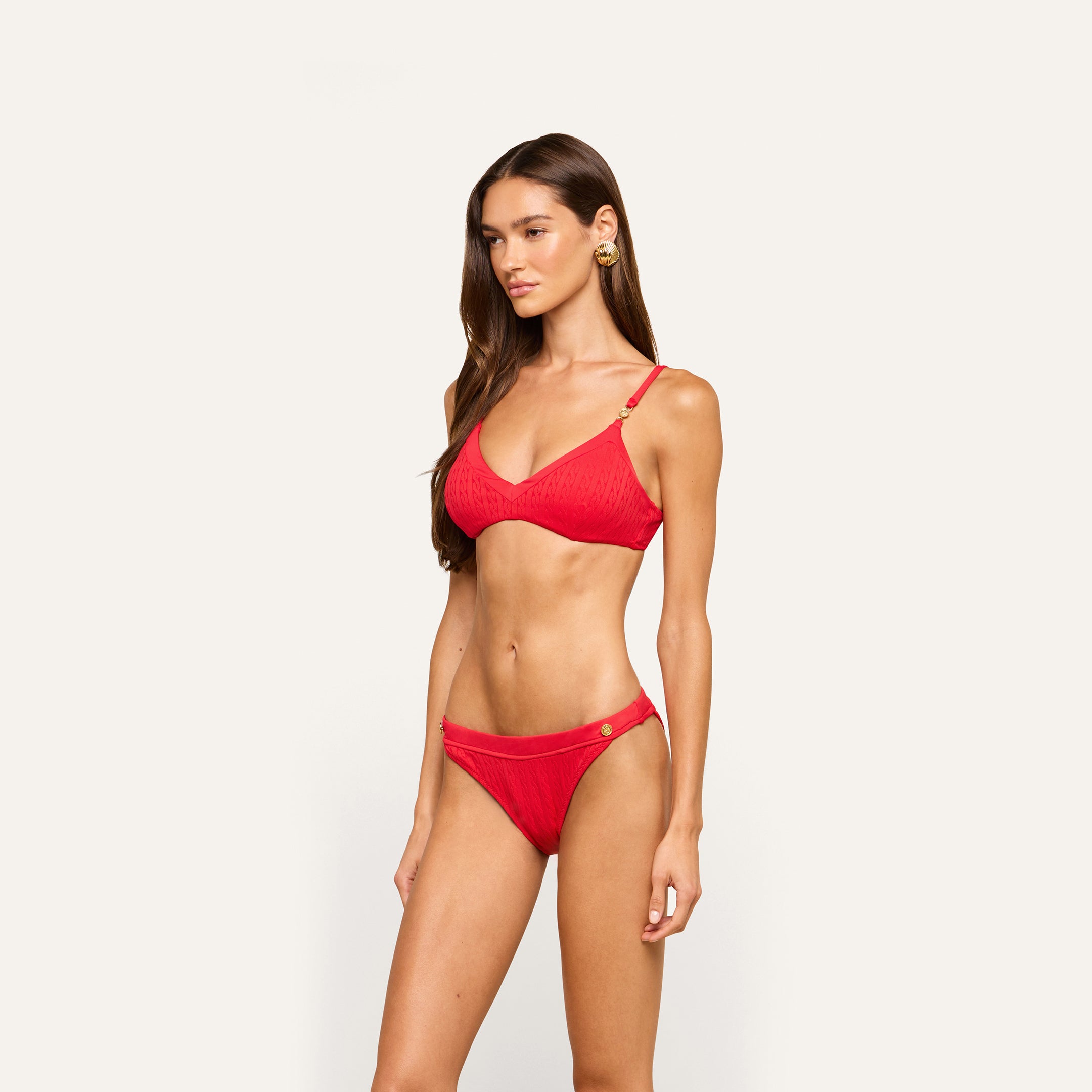 A woman with long brown hair wears the Jean Bikini Bottom in red, crafted from textured swim fabric, paired with gold earrings, standing against a plain off-white background.