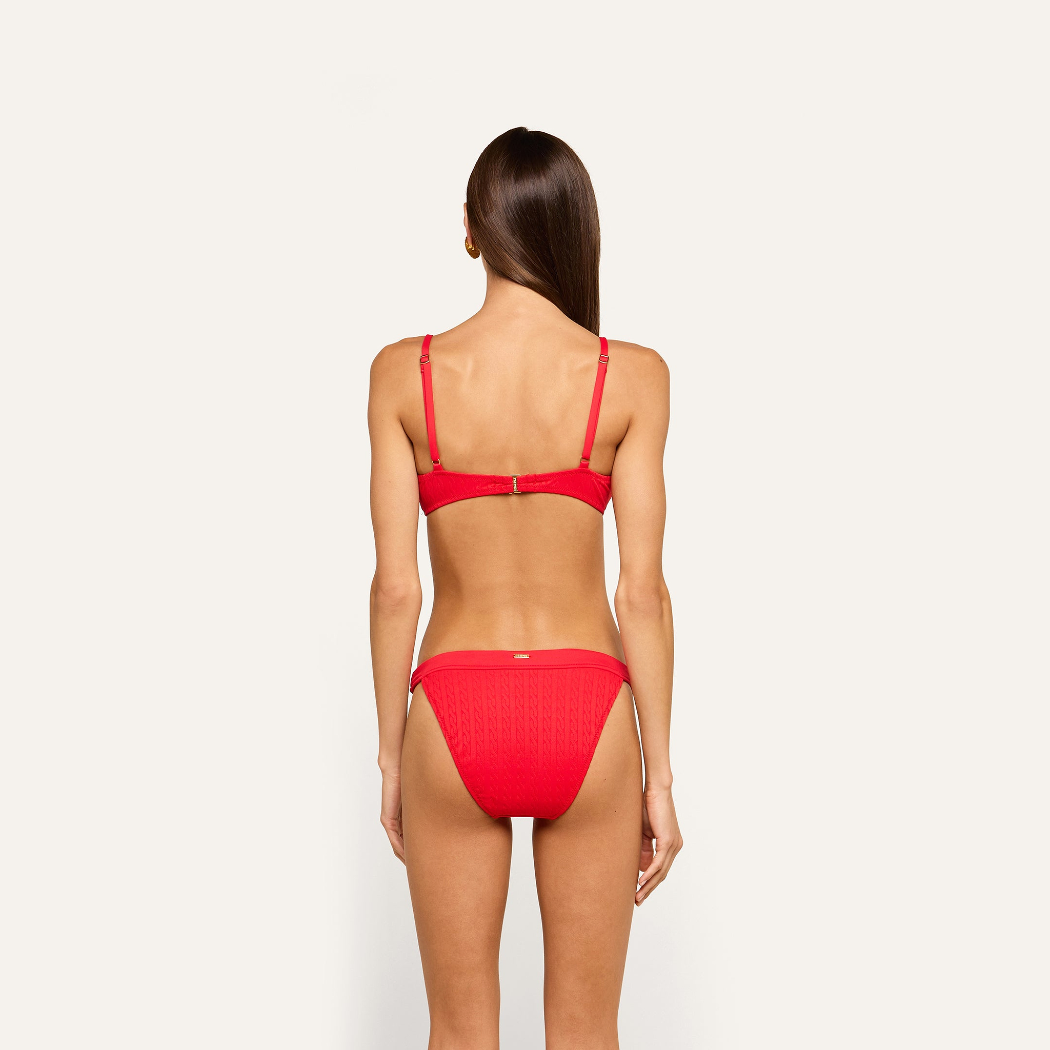 A woman with long straight hair stands facing away, wearing a red Jean Bikini Bottom crafted from textured swim fabric with adjustable straps and a high-leg silhouette, set against a plain light background.