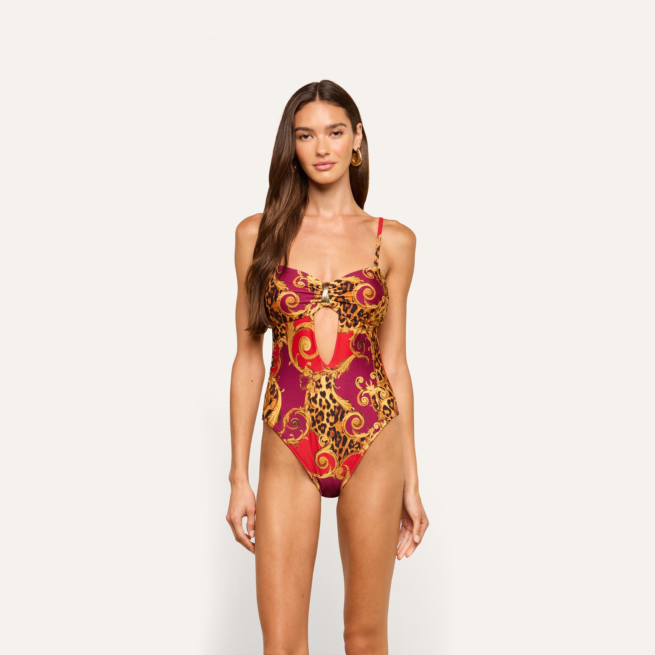 A woman stands against a plain light background, wearing the Emilia Bandeau One-Piece Swimsuit—a red and yellow patterned bandeau with a keyhole cutout, perfect for resort swimwear.