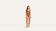 A woman with long hair wears a red and gold patterned one-piece swimsuit while standing barefoot against a plain white background, exuding confidence and elegance.