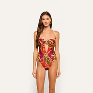 A woman wearing the Emilia Bandeau One-Piece Swimsuit, featuring a strapless design, red and gold pattern, and a front cutout, stands against a plain light background.