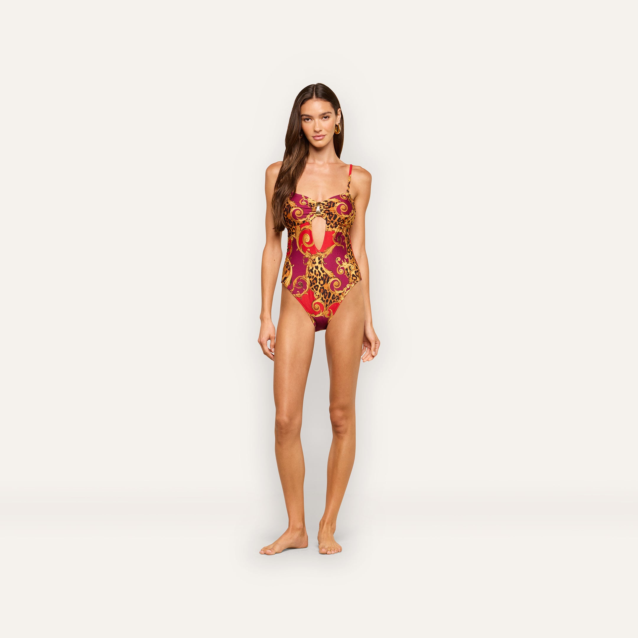 A woman stands barefoot in the Emilia Bandeau One-Piece Swimsuit, featuring gold ornate patterns on a red base, against a plain light background.