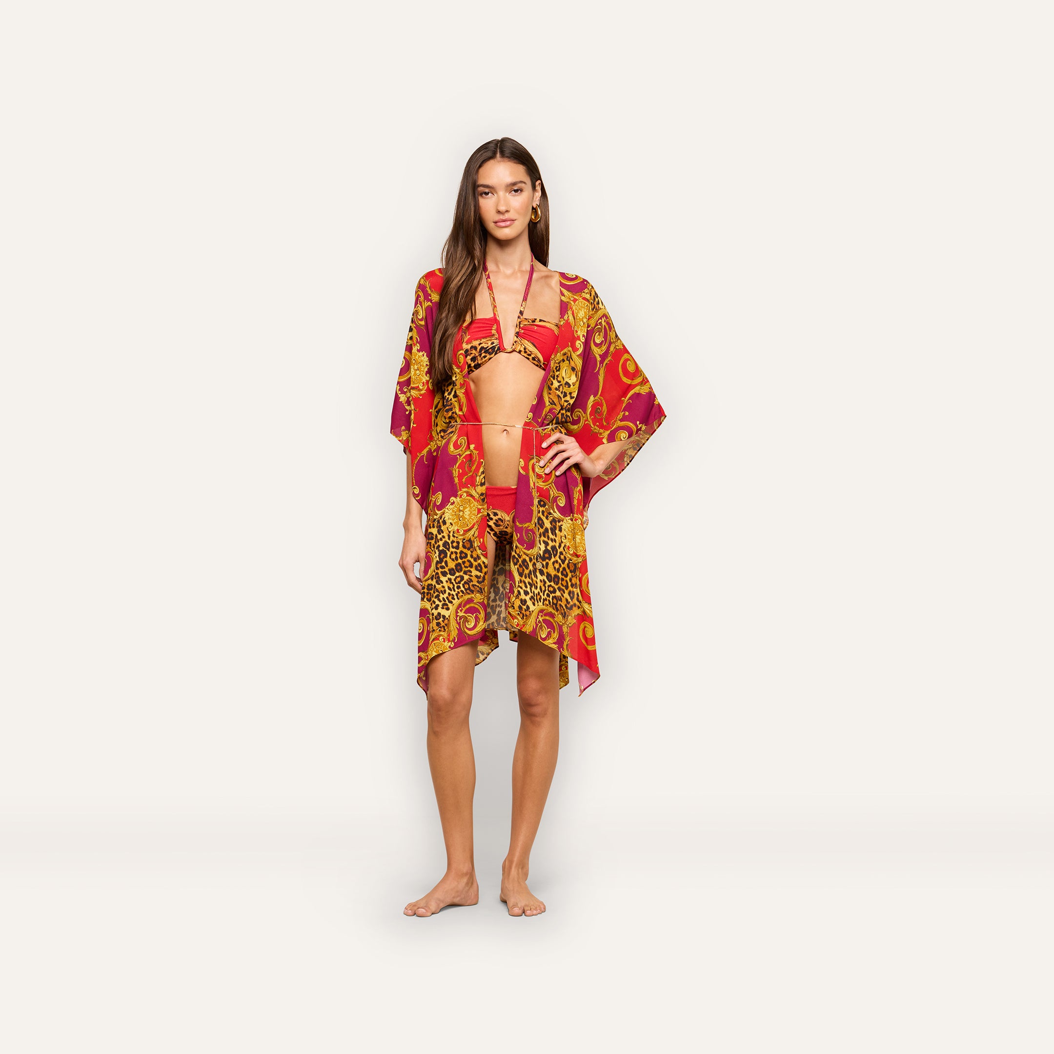 A woman stands barefoot against a plain off-white background, wearing a red and yellow patterned bikini with the Kara Cover-Up Kimono and a gold chain belt.