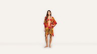 A woman stands barefoot wearing a red and gold patterned bikini and matching cover-up against a plain white background, exuding effortless elegance.