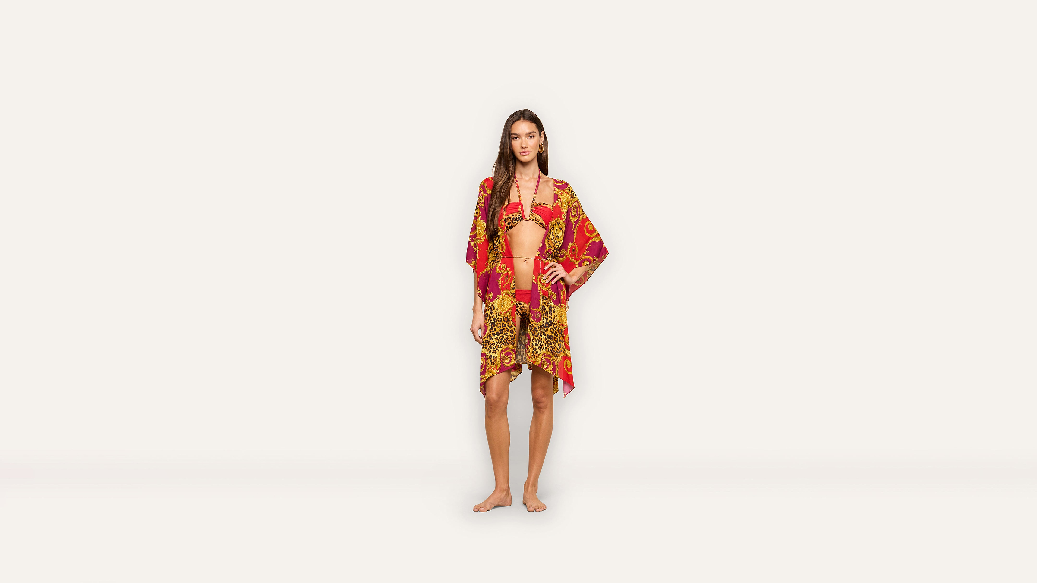 A woman stands barefoot wearing a red and gold patterned bikini and matching cover-up against a plain white background, exuding effortless elegance.