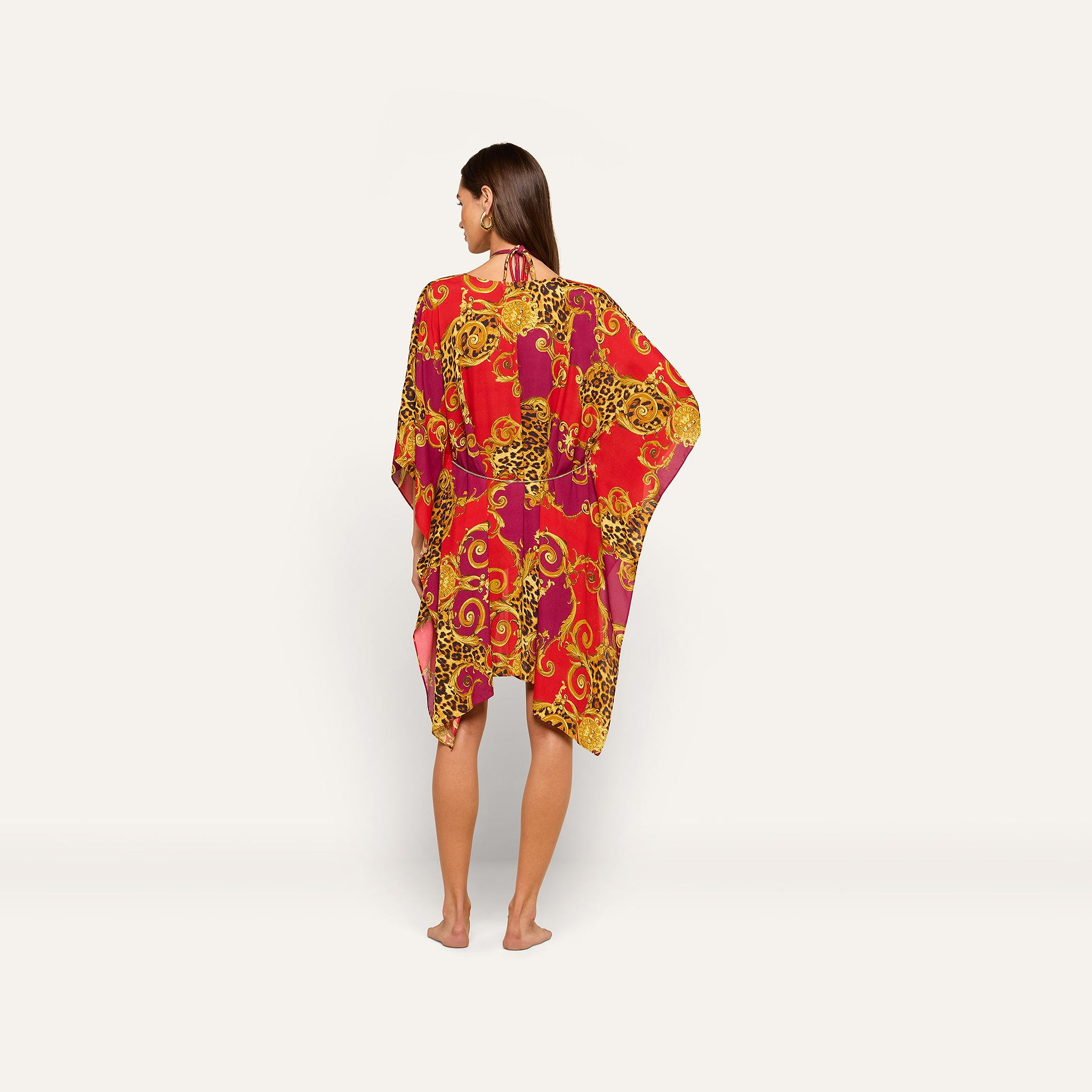 A woman stands barefoot facing away, wearing the Kara Cover-Up Kimono—a vibrant red, pink, and yellow patterned kimono-style dress with loose sleeves and a gold chain belt—set against a plain white background.