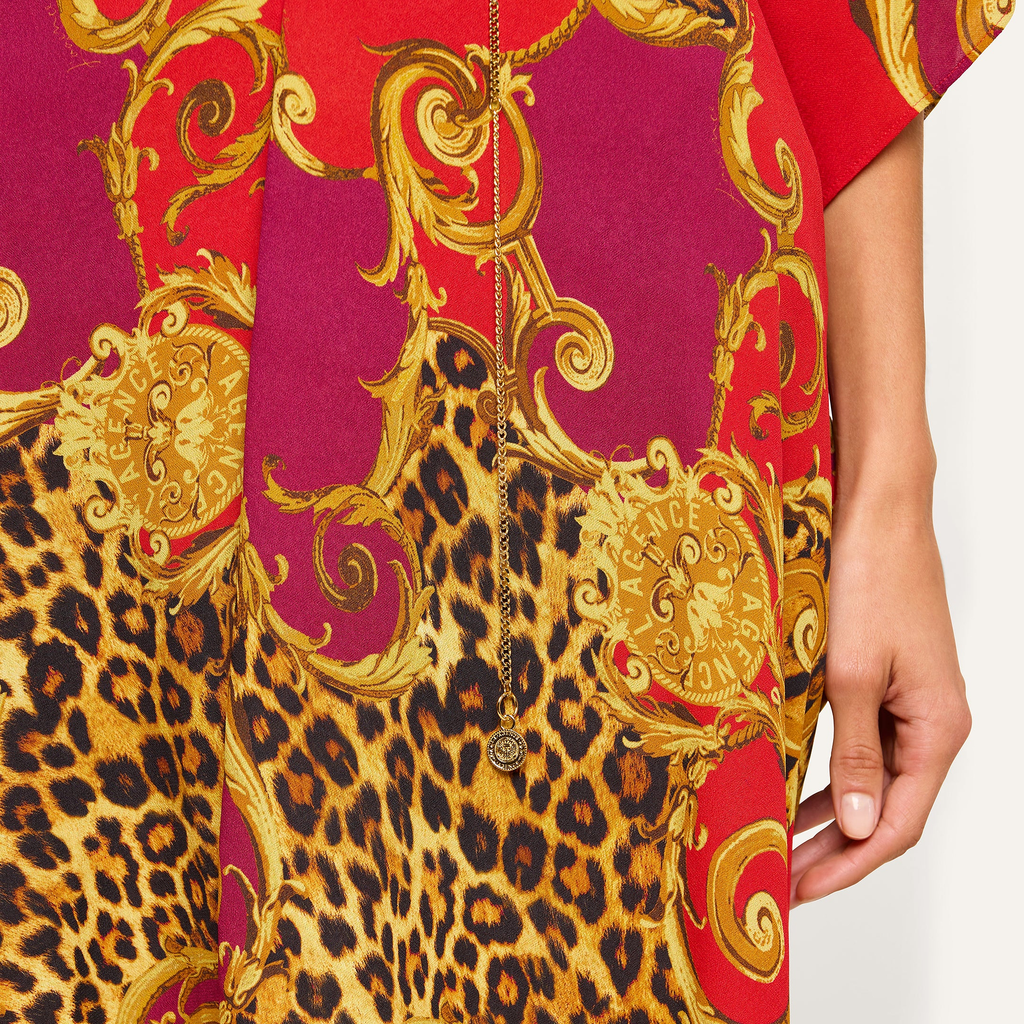A close-up of the Kara Cover-Up Kimono, showcasing its vibrant red and gold baroque design with leopard print accents. Styled as a striking leopard print cover-up, it is complemented by a delicate gold necklace with a small pendant resting on the patterned fabric near a person’s hand.