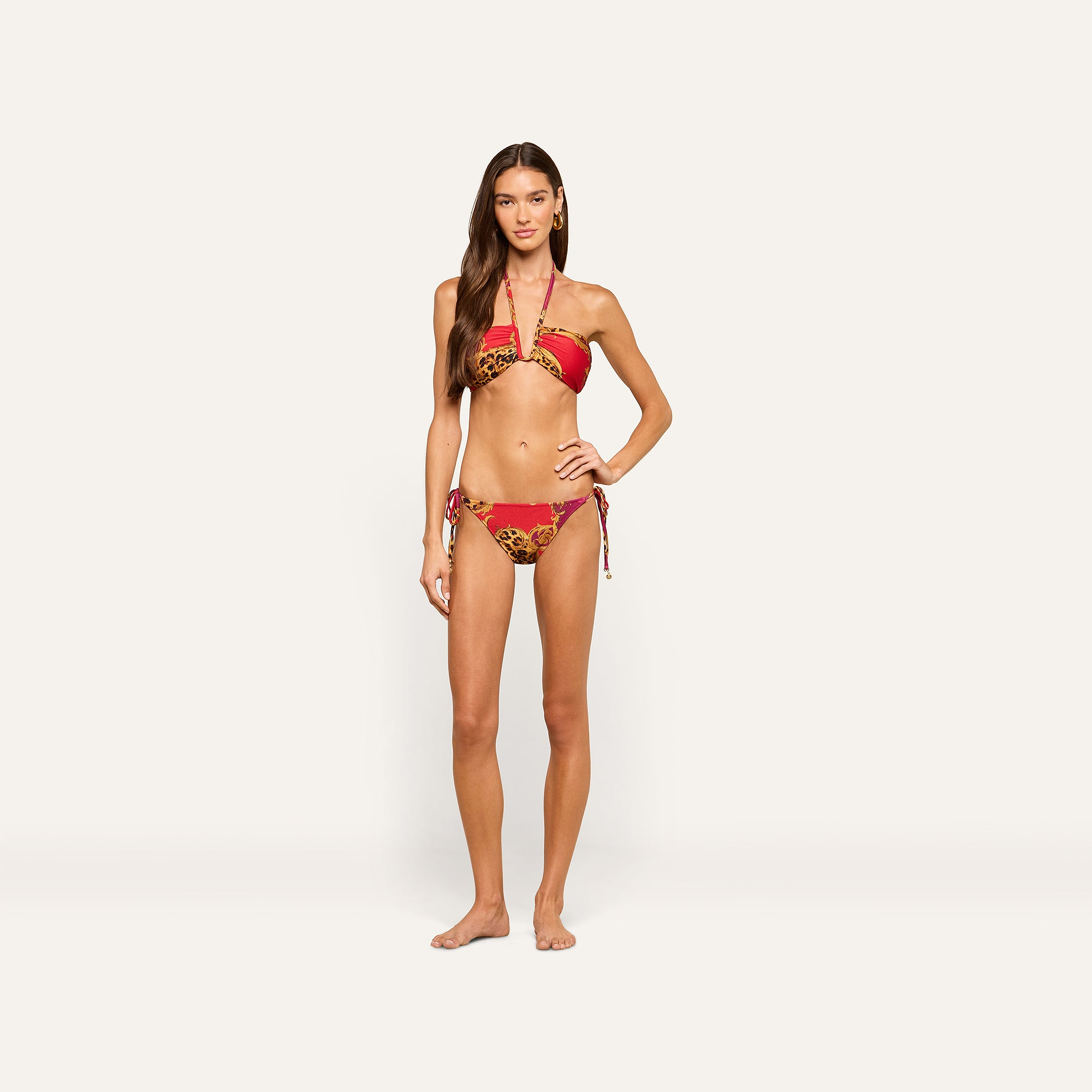 A woman poses against a plain background wearing the Britt Tie-Side Bikini Bottom in a gold leopard print, standing with one hand on her hip and the other relaxed at her side.