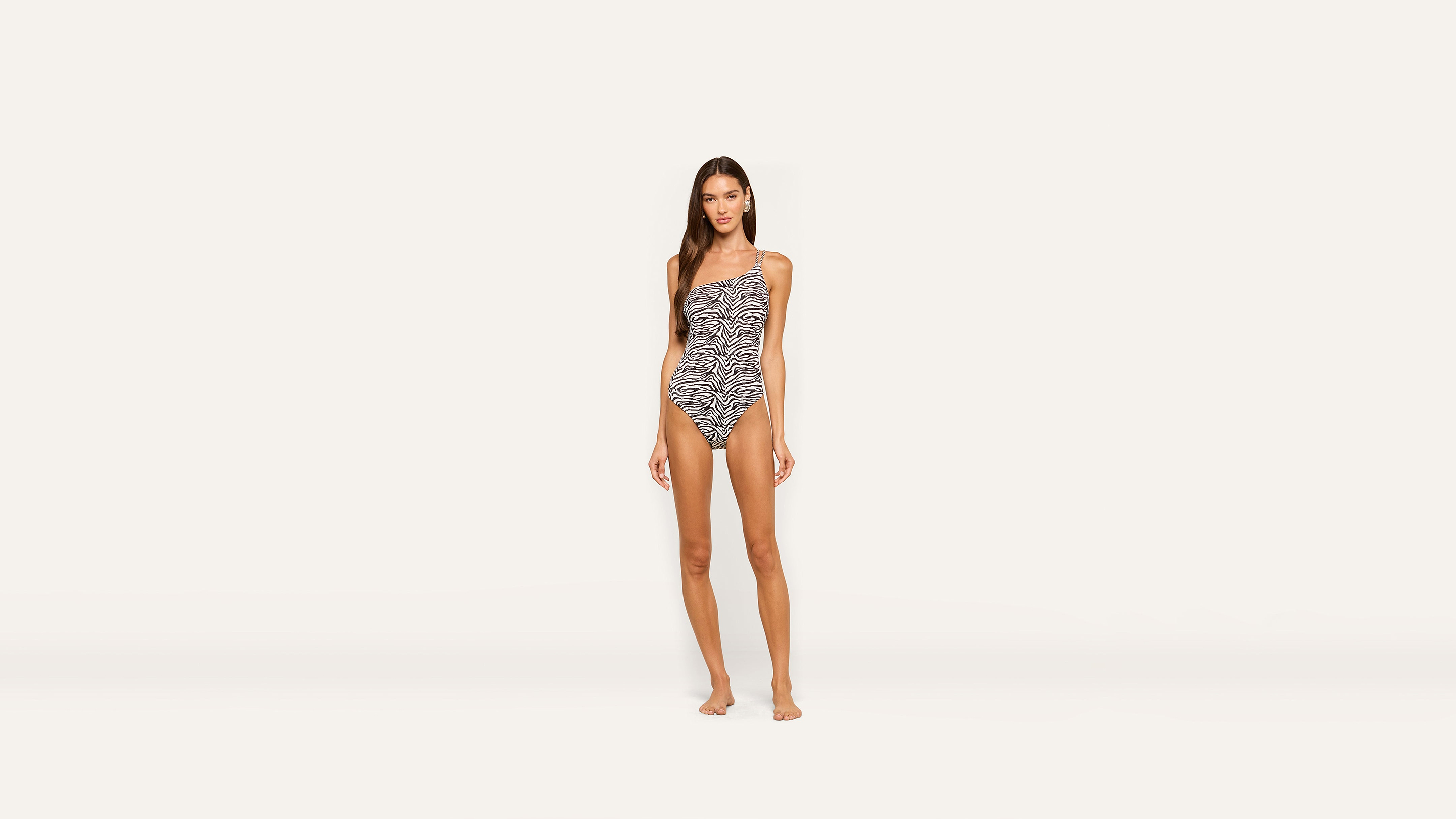 A woman stands barefoot in a stylish one-shoulder zebra print swimsuit against a plain, light background.