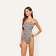 A woman with long brown hair poses against a plain light background, wearing the Karmin One-Piece Swimsuit—a high-cut, one-shoulder zebra print design with a side strap.
