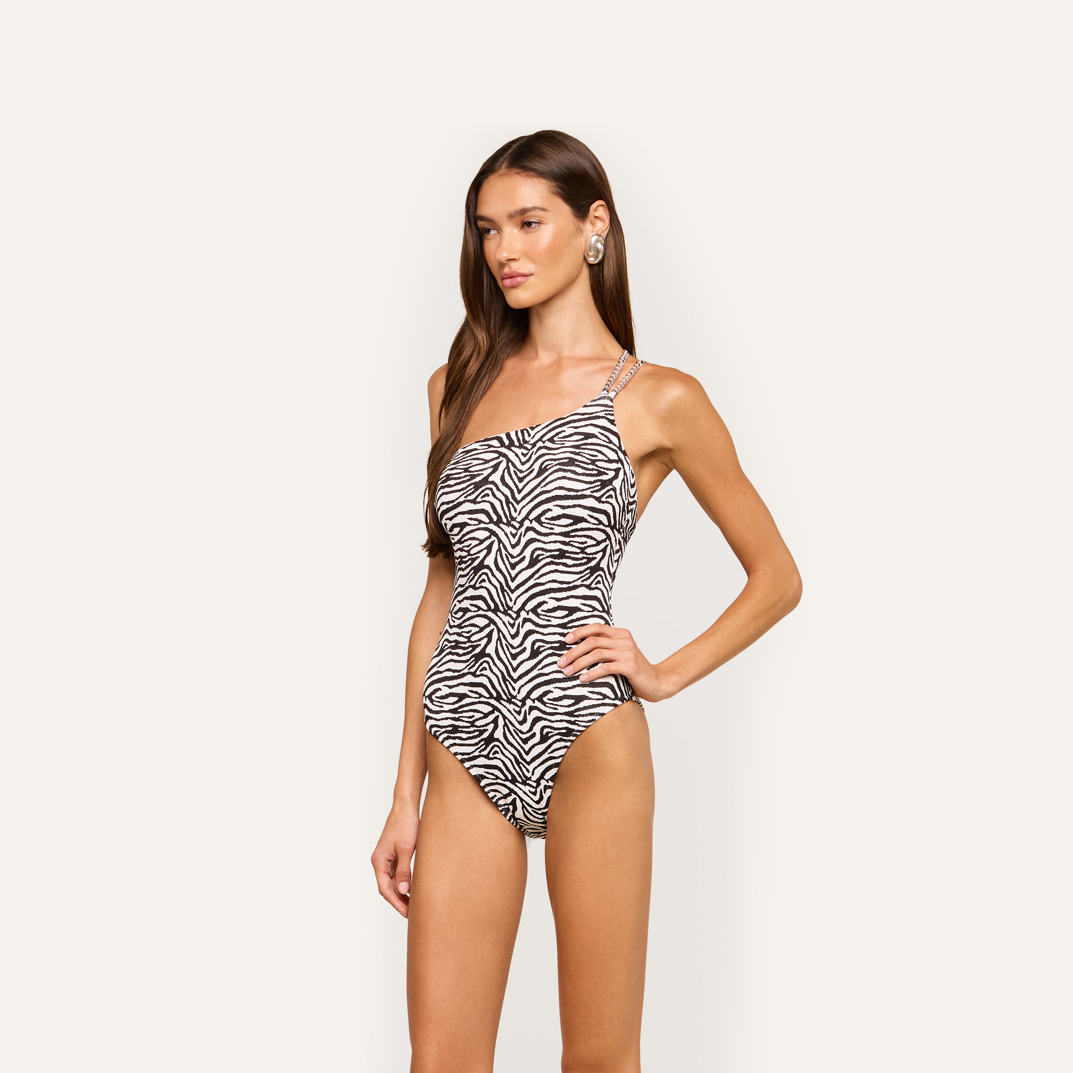 A woman with long brown hair poses against a plain light background, wearing the Karmin One-Piece Swimsuit—a high-cut, one-shoulder zebra print design with a side strap.