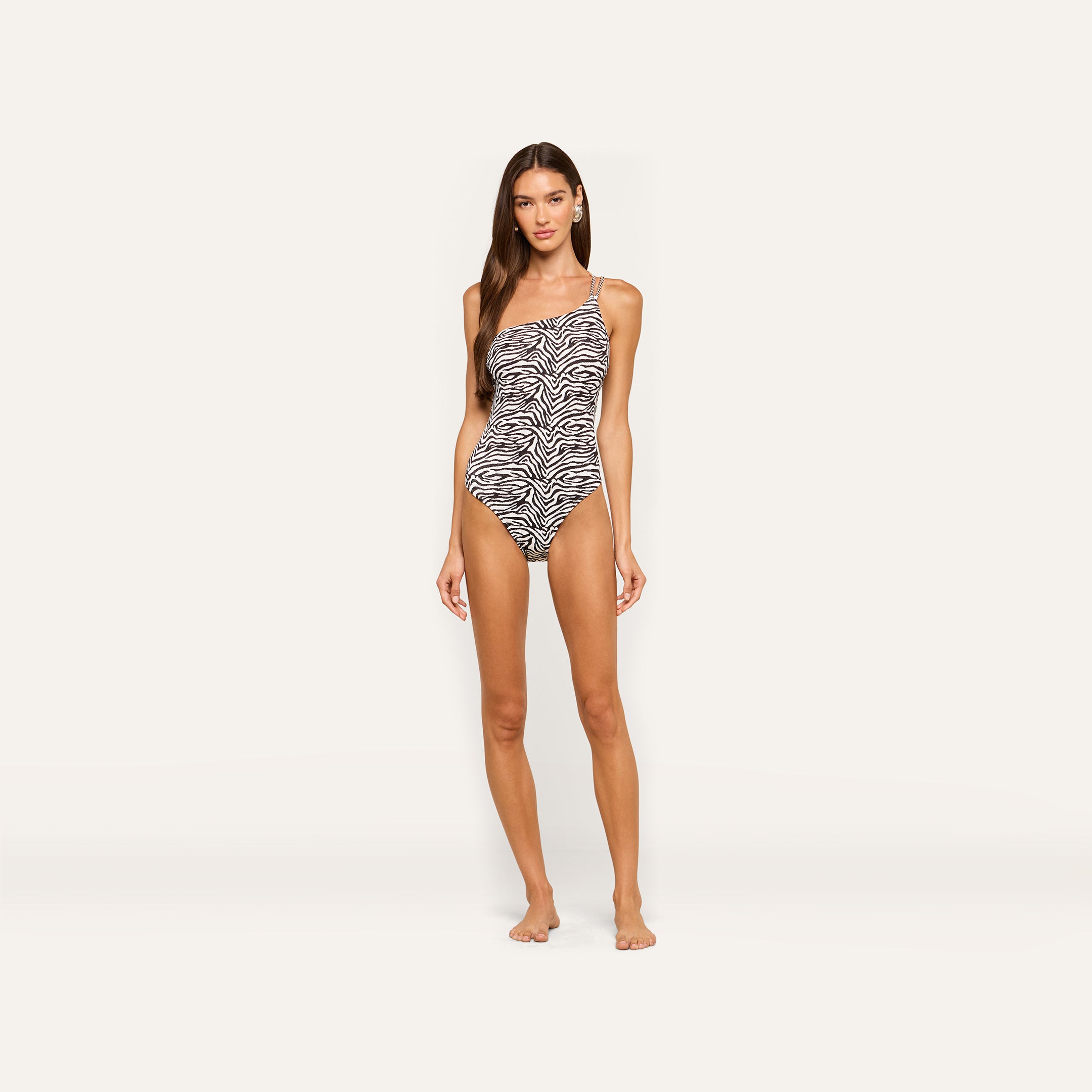 Woman stands barefoot in the Karmin One-Piece Swimsuit, featuring a zebra print, one-shoulder design with thin straps against a plain light background.