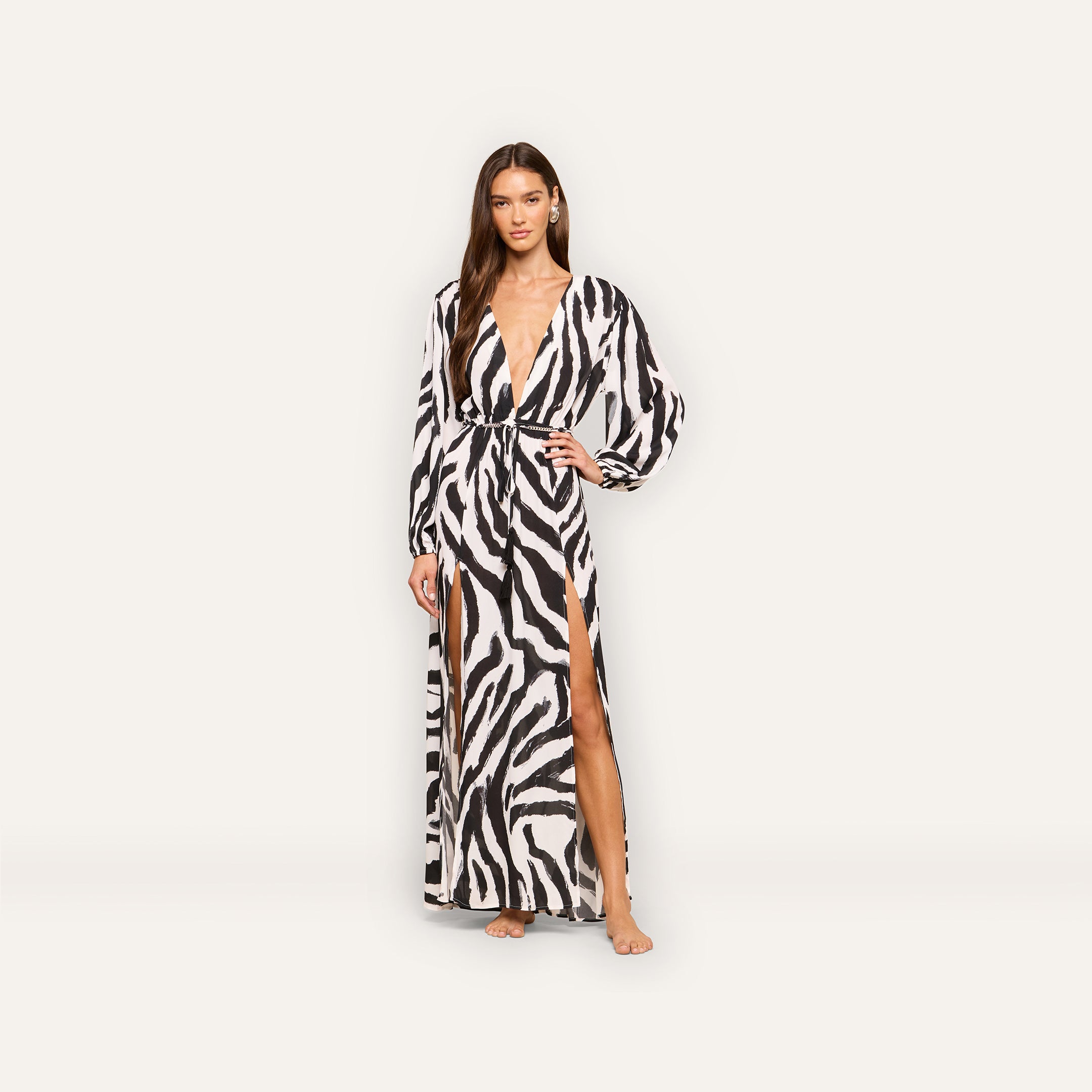 A woman stands barefoot against a plain light background, wearing the Sara Maxi Cover-Up Dress—a long zebra print dress with long sleeves, a deep V-neckline, and a high side slit.