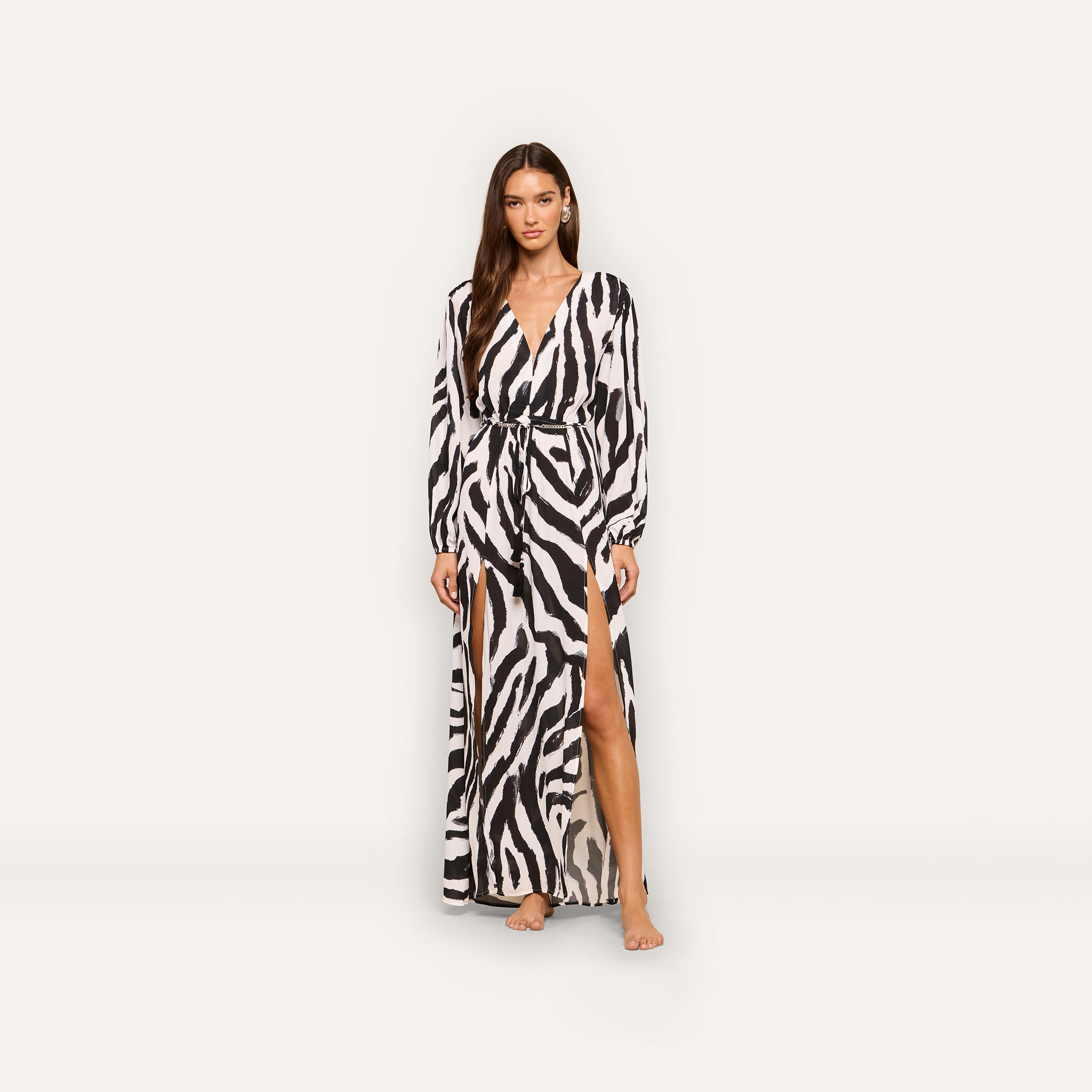A woman stands barefoot in the Sara Maxi Cover-Up Dress, which features a zebra print, long sleeves, a deep V-neck, a belted waist, and high front slits, set against a plain white background.